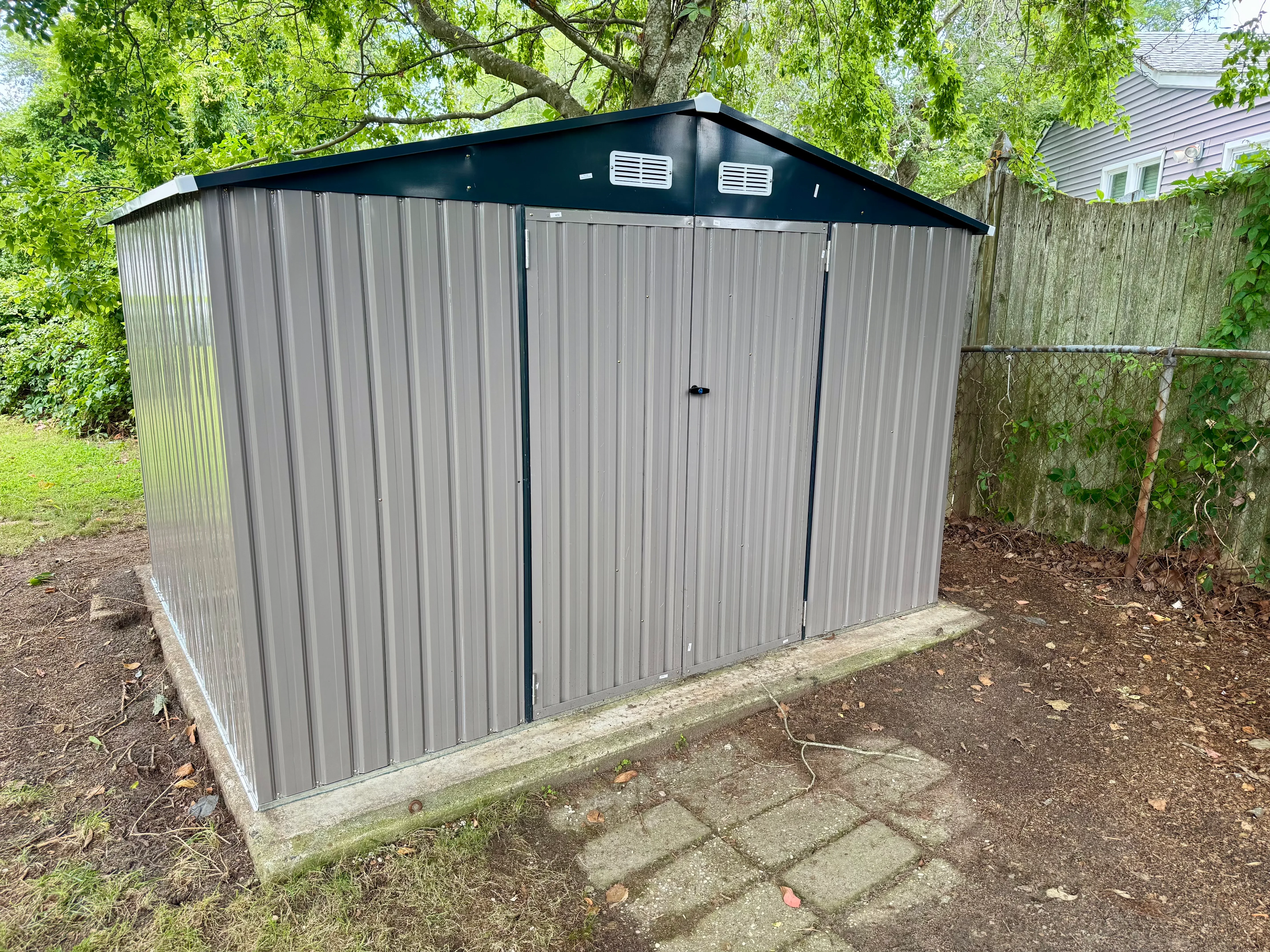 Gray corrugated metal storage shed with dark green roof and double doors on concrete foundation